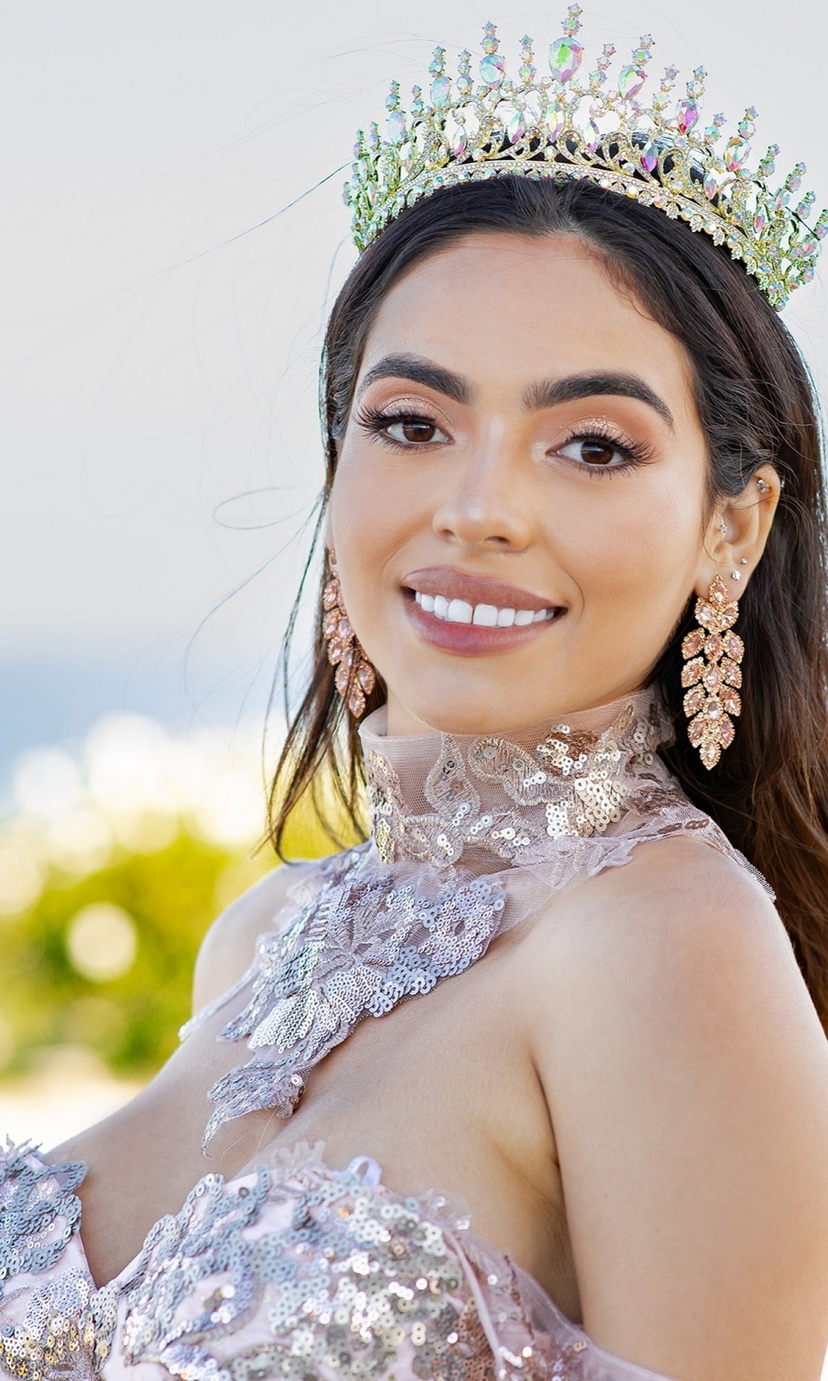 A woman smiles outdoors, wearing the Quinceanera Ball Gown 8064J—an ornate off-the-shoulder dress with lace embroidery—paired with a jeweled crown and sparkly earrings.